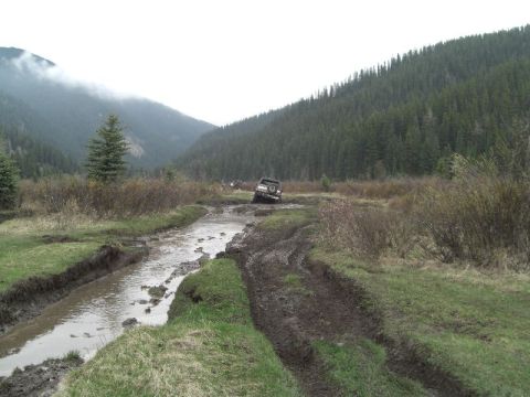 1995 Ford Bronco - A 1995 Ford Bronco tackles a challenging muddy off-road path surrounded by dense forest and misty mountains.