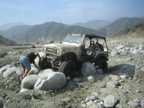 1987 Suzuki Samurai - A person inspects the front tire of a lifted Suzuki Samurai as it maneuvers through a rocky, dry riverbed.
