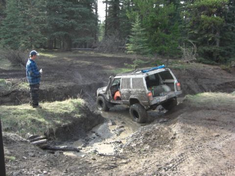 1995 Ford Bronco - A person watches a muddy SUV navigate a challenging off-road course in a wooded area.