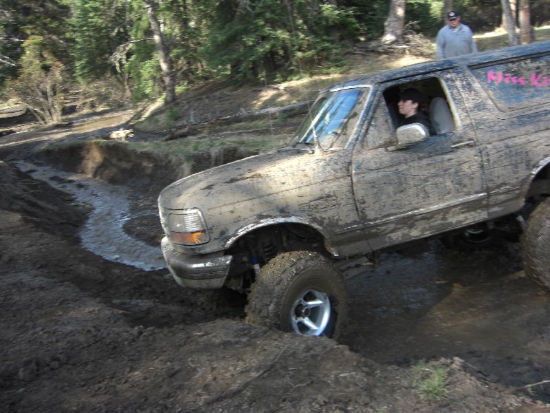 Muddy 1995 Ford Bronco driving through a muddy ditch with trees in the background. A person is in the driver's seat