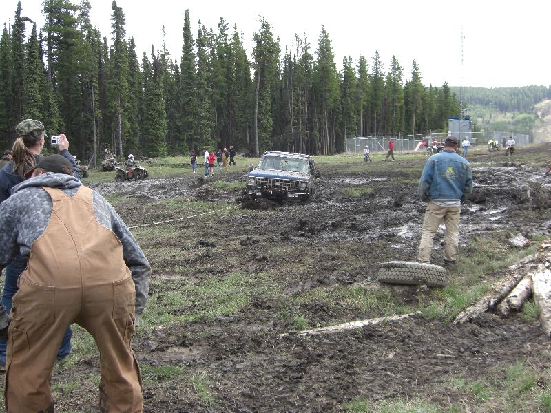 Off-road vehicle stuck in mud pit with trees in background. Several people are watching