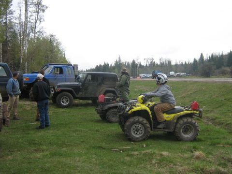 1995 Ford Bronco - Several off-road vehicles, including ATVs and trucks, are gathered on a grassy field with trees and a distant road.