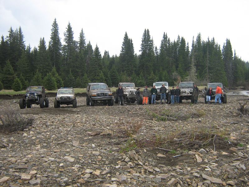 Brown SUV with a group of people and other off-road vehicles in a rocky, wooded area