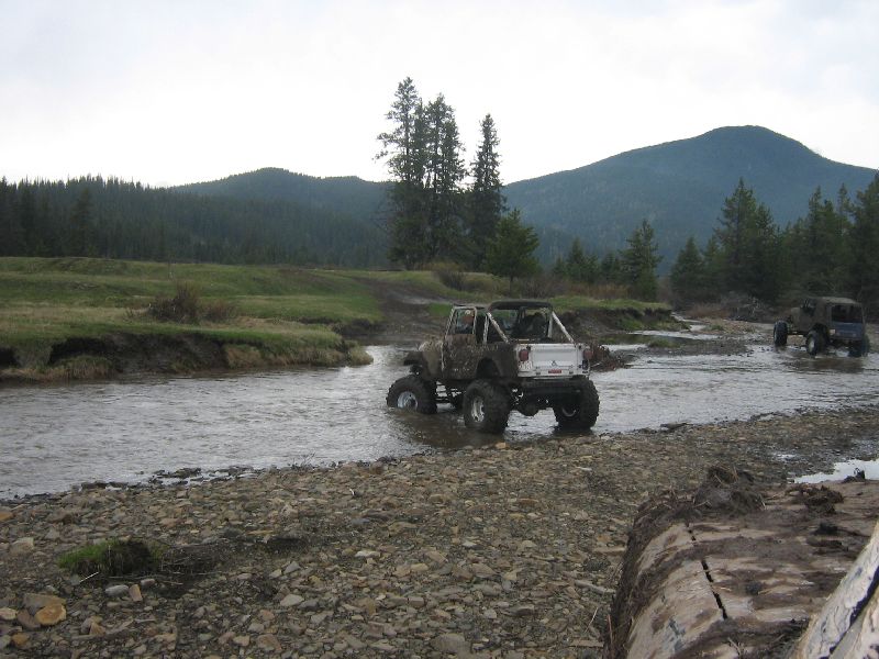 Two modified off-road vehicles drive through a shallow stream with wooded hills in the background