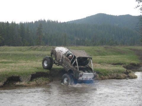 1995 Ford Bronco - An off-road vehicle navigates a muddy stream with a wooded hillside in the background.
