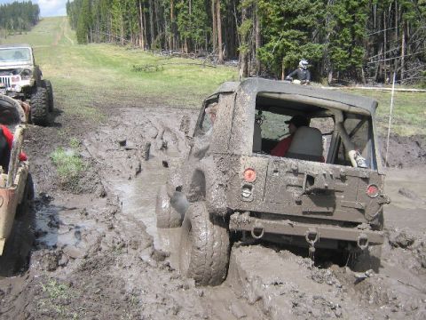 1995 Ford Bronco - Several off-road vehicles navigate a challenging muddy terrain with dense forest and a grassy slope.