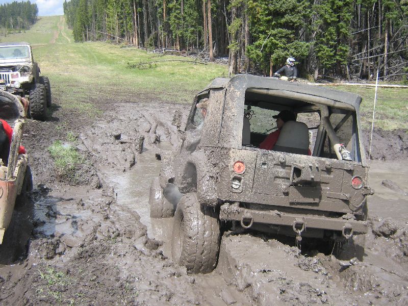 Jeep Wrangler stuck in deep mud, another Jeep visible to the left, wooded area and grassy hill in background