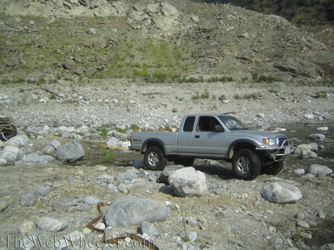 1987 Suzuki Samurai - A silver pickup truck navigates a rocky, dry riverbed, equipped for off-road conditions.