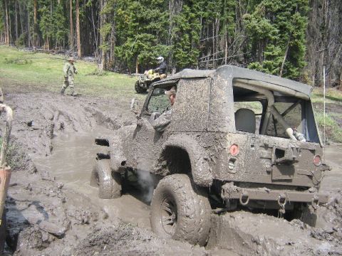 1995 Ford Bronco - A heavily mud-covered Jeep Wrangler struggles in a deep mud pit, with an ATV visible in the background.