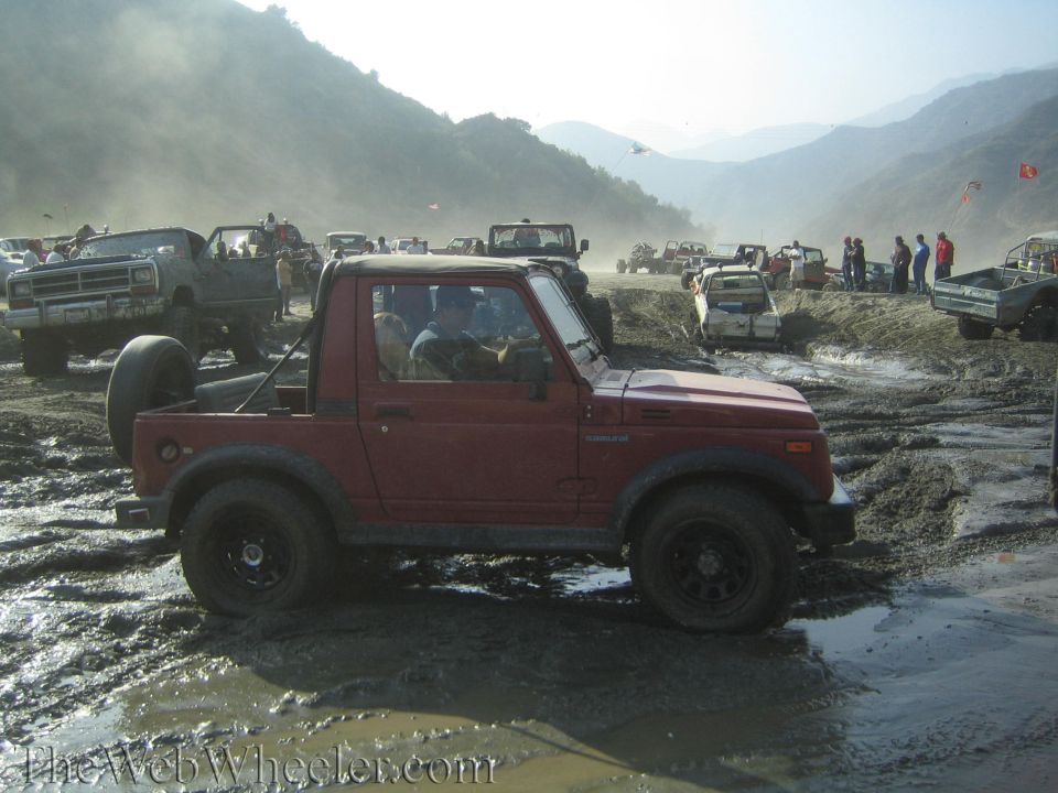 Red 1987 Suzuki Samurai driving through mud with other vehicles in background