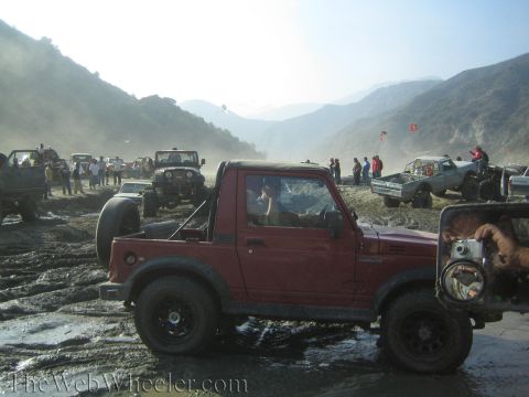 1987 Suzuki Samurai - A group of off-road vehicles are gathered in a muddy, hilly area under a hazy sky.
