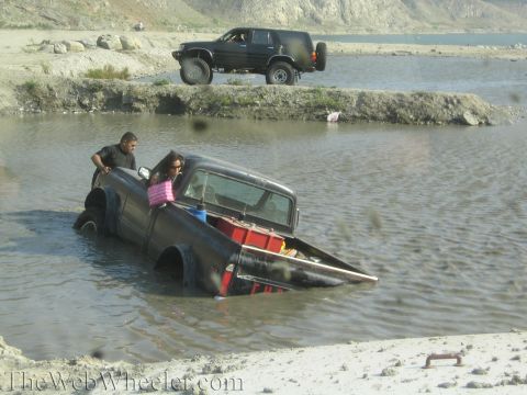 1987 Suzuki Samurai - A black pickup truck is partially submerged in water, with two people inside.