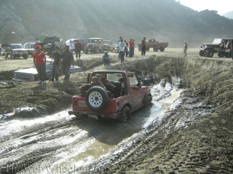 1987 Suzuki Samurai - A red 1987 Suzuki Samurai navigates a muddy obstacle course with other vehicles and spectators present.