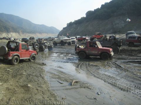 1987 Suzuki Samurai - Several off-road vehicles, including a red 1987 Suzuki Samurai, navigate a muddy terrain under a bright sky.