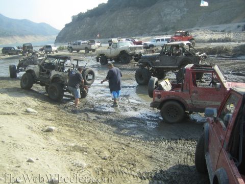 1987 Suzuki Samurai - Off-road vehicles gathered in a muddy area, with a body of water and hills in the background.