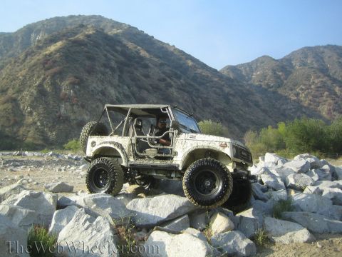 1987 Suzuki Samurai - A white Suzuki Samurai is shown navigating a rocky terrain, with a driver visible inside the open-top vehicle.