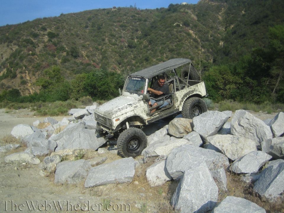 White 1987 Suzuki Samurai climbing over large rocks on a dirt trail with a hilly, vegetated background