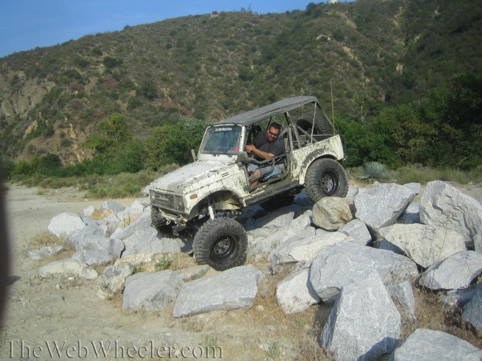 White 1987 Suzuki Samurai climbing over large rocks on a dirt path with a man driving