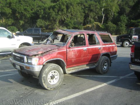 1987 Suzuki Samurai - This damaged red Toyota SUV is parked in a lot with other vehicles and trees in the background.