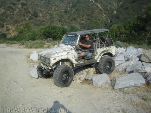 1987 Suzuki Samurai - Mud-covered 1987 Suzuki Samurai tackling a rocky off-road course with a driver at the wheel.