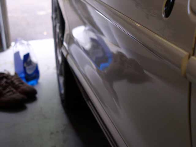 Close-up of a car's side panel and wheel well, with shoes and a bottle of fluid in the background