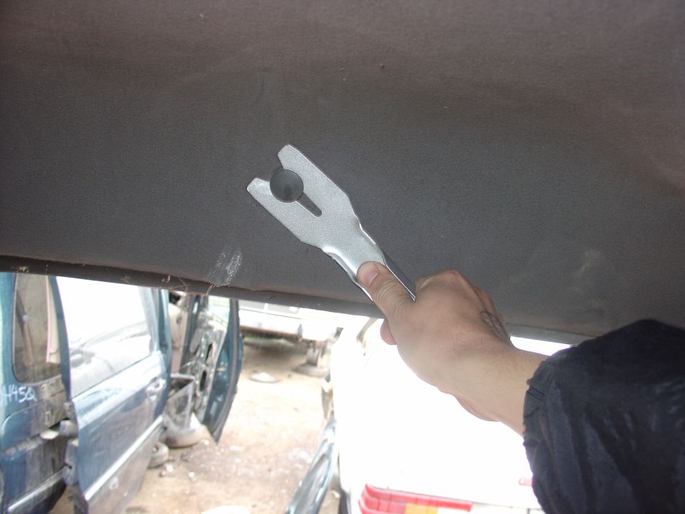 Hand holding a metal trim removal tool against a car's interior headliner. Other vehicles visible in background