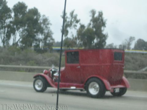 1990 Ford Bronco - dark red tall-cabin hot rod with exposed engine and wide rear tires drives on a freeway past trees.