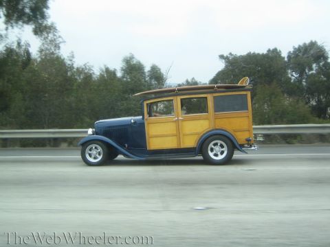 1990 Ford Bronco - blue vintage woodie station wagon carrying a surfboard cruises a freeway with chrome wheels.