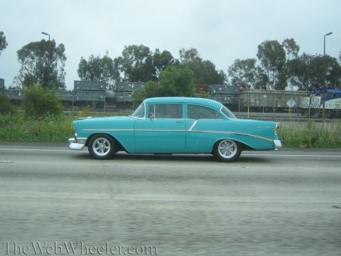 1990 Ford Bronco - teal 1956-style two-door classic sedan with chrome trim drives a highway past a rail yard.