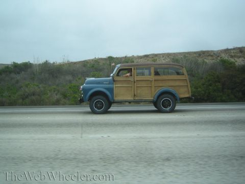 1990 Ford Bronco - blue vintage woodie wagon with wood-paneled sides travels a highway with brush-covered hills behind.
