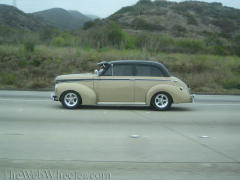 1990 Ford Bronco - tan 1940s-style convertible street rod with a black soft top cruises a freeway by green hills.