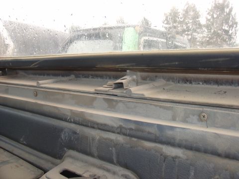 1988 Ford Bronco - Dust covers the interior trim and window mechanism of a vehicle, with a glimpse of the outside world through the rain-streaked glass.