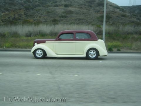 1990 Ford Bronco - cream-bodied 1930s coupe street rod with maroon top drives a freeway beside grassy hills.