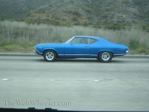 1990 Ford Bronco - blue 1968-style two-door muscle coupe with chrome wheels travels a highway past a brushy hillside.