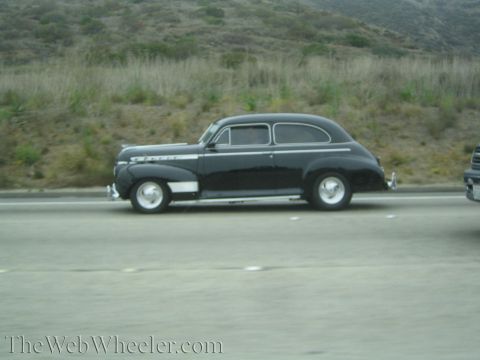 1990 Ford Bronco - black 1940s-style two-door sedan with chrome trim and whitewall-style wheels cruises a highway.