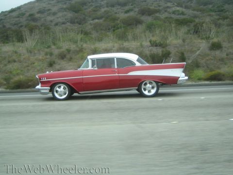 1990 Ford Bronco - red and white 1957-style Chevrolet two-door hardtop with tailfins drives a highway by green hills.