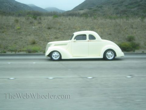 1990 Ford Bronco - cream-colored 1937-style coupe street rod with chrome wheels cruises a freeway beside grassy hills.