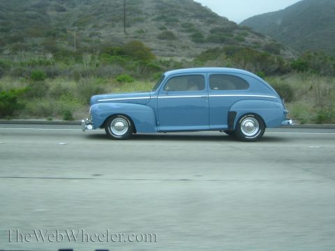 1990 Ford Bronco - blue 1940s-style two-door sedan street rod with chrome wheels travels a freeway by green hills.