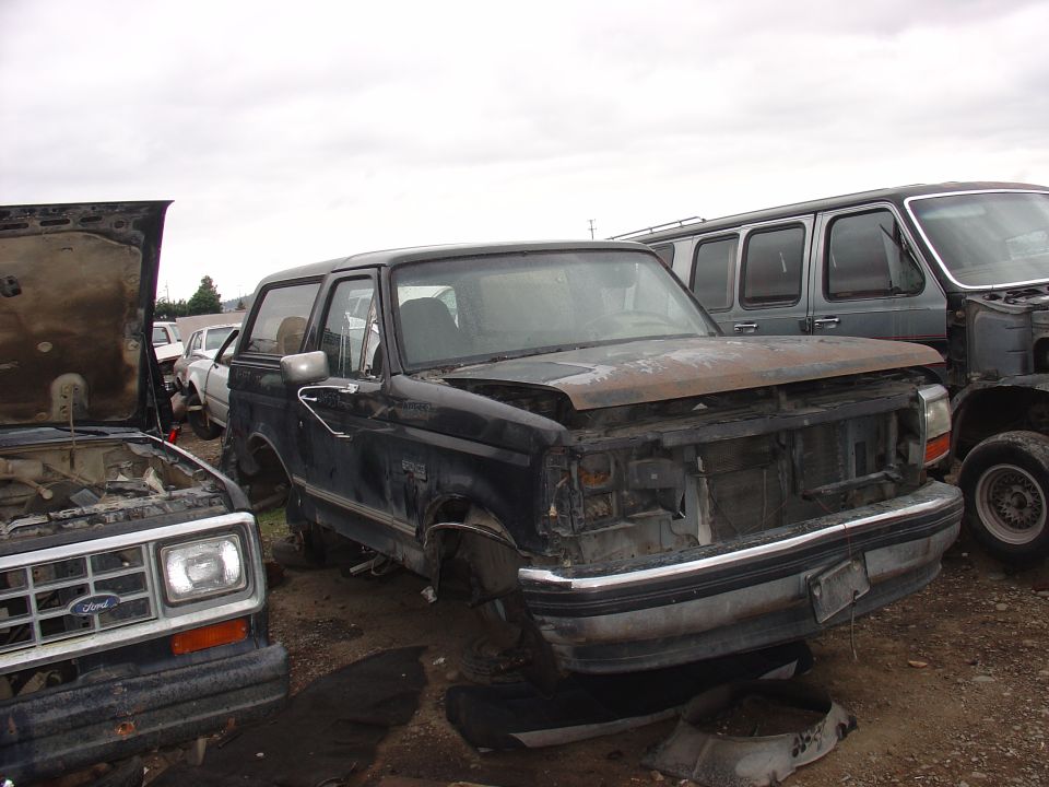 Black Ford Bronco in junkyard, missing grille and hood. Another vehicle is in the foreground with hood open