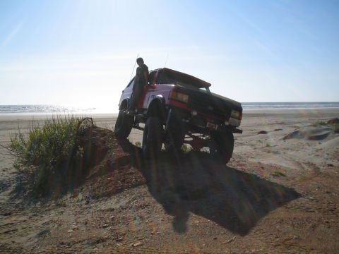 1989 Ford Bronco - A person sits on the side of a heavily modified SUV parked on a sand dune overlooking the ocean.