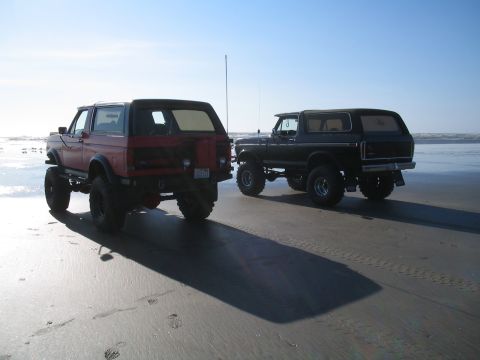1989 Ford Bronco - Two lifted Ford Broncos parked on the sand near the ocean under a clear sky.