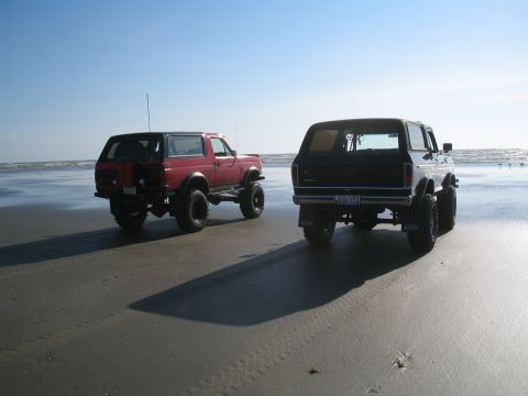 1989 Ford Bronco - Two Broncos are parked on the beach under a clear blue sky.