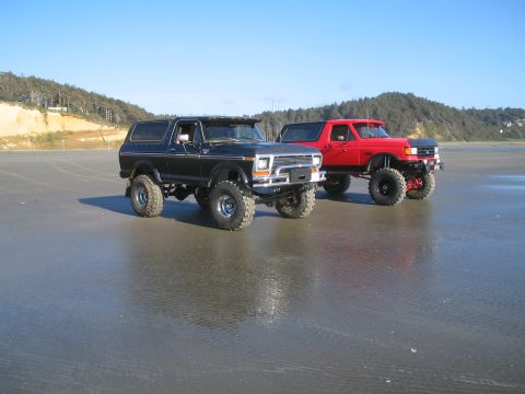 1989 Ford Bronco - Two lifted Ford Broncos are parked on a wet beach under a clear blue sky.