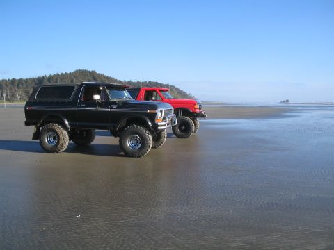 1989 Ford Bronco - A black 1978 Ford Bronco and a red pickup truck sit on a sandy beach with the ocean in the background.