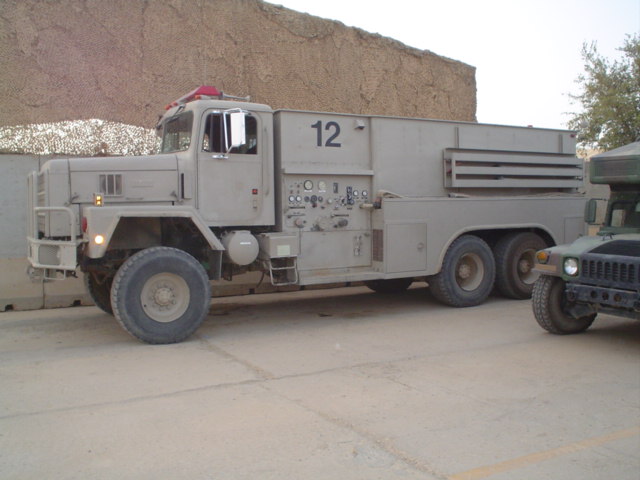 Tan military truck with a water tank body and the number 12 on its side, parked next to a Humvee