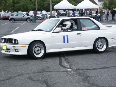 2001 Mercury Grand Marquis - A white BMW E30 M3 coupe, identifiable by its distinctive body style and racing stripes, participates in an event. The driver is wearing a…