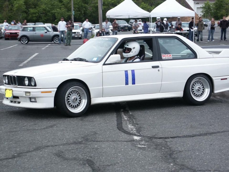 White BMW E30 M3 coupe with blue racing stripes and BBS wheels, driver in helmet