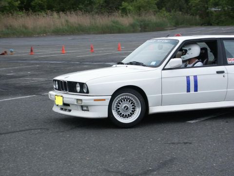 2001 Mercury Grand Marquis - White BMW E30 M3 with blue racing stripes on driver's door, on asphalt track with orange cones in background.
