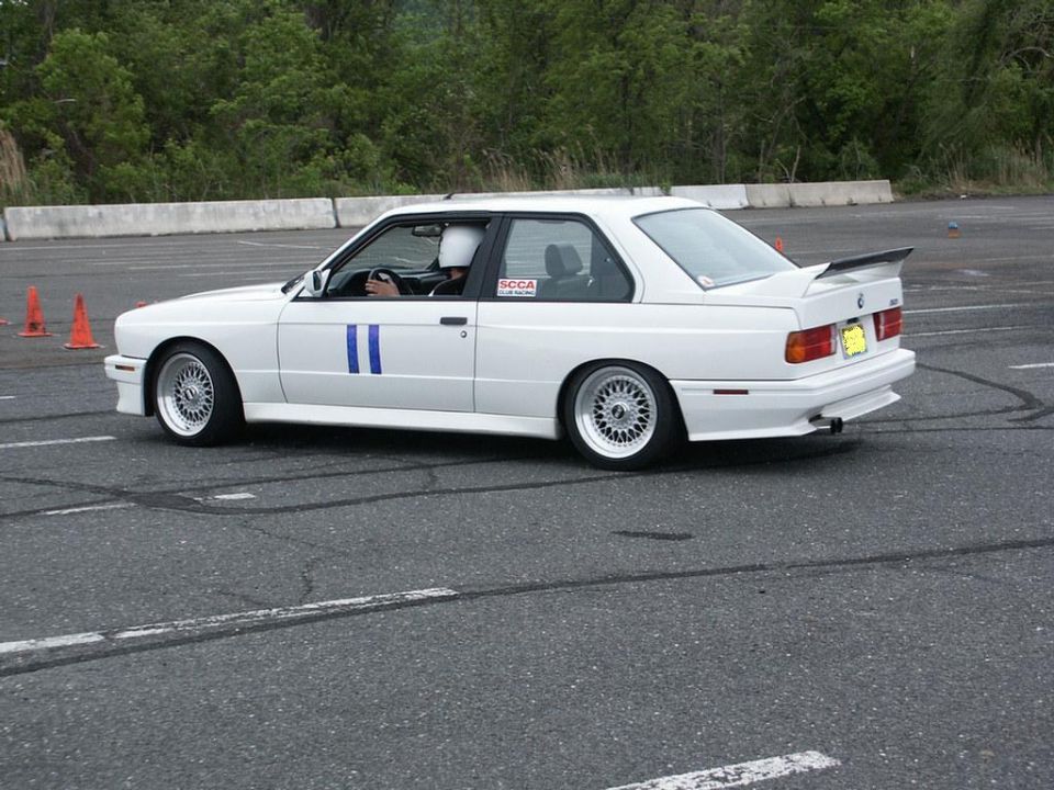 White BMW E30 M3 with blue racing stripes on side, spoiler, and BBS wheels on a track