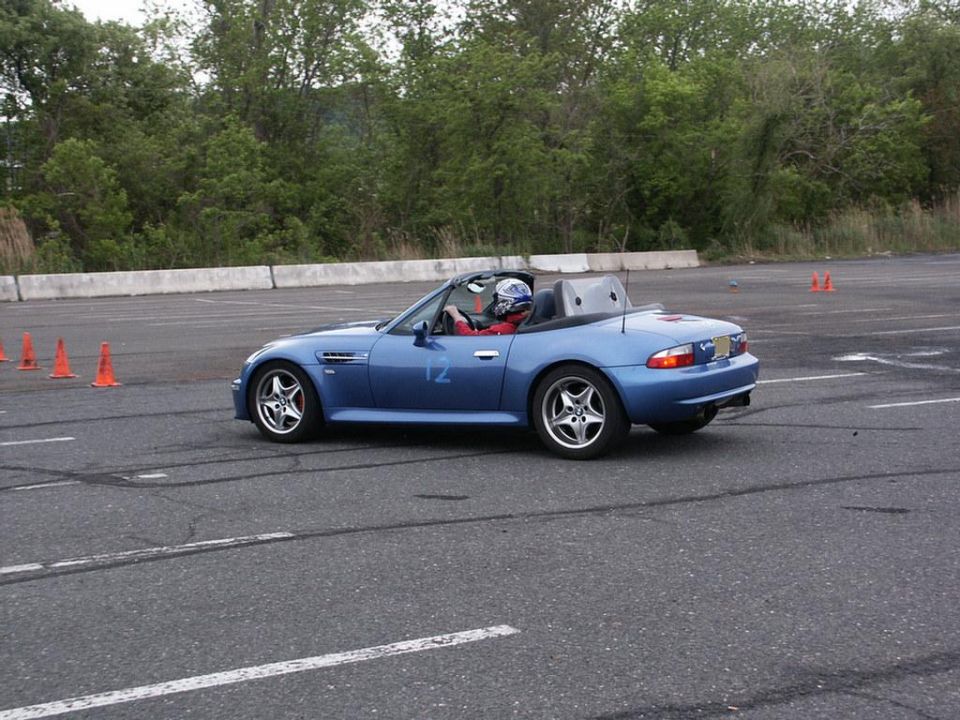 Blue convertible sports car with racing number 12 on the door, driven by a person in a helmet, on a paved track with orange cones